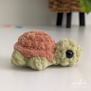May include: A small crochet turtle toy made from soft, fuzzy yarn. The turtle features a dusty rose-colored shell and a light sage green head, body, and flippers. It has a single visible black plastic safety eye on the side of its head.