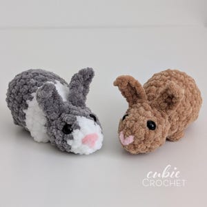 Op de afbeelding: Twee gehaakte pluche konijntjes. E&eacute;n is grijs met witte en zwarte accenten en een roze neus. De andere is bruin met een roze neus. De konijntjes liggen. De tekst "cubie CROCHET" staat in de rechter benedenhoek.