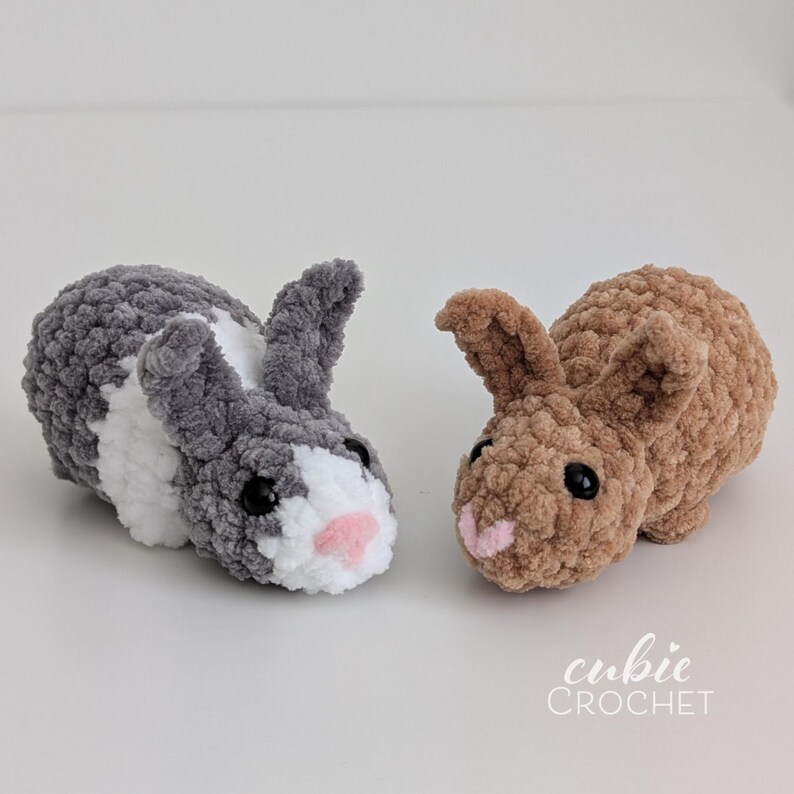 Op de afbeelding: Twee gehaakte pluche konijntjes. E&eacute;n is grijs met witte en zwarte accenten en een roze neus. De andere is bruin met een roze neus. De konijntjes liggen. De tekst "cubie CROCHET" staat in de rechter benedenhoek.
