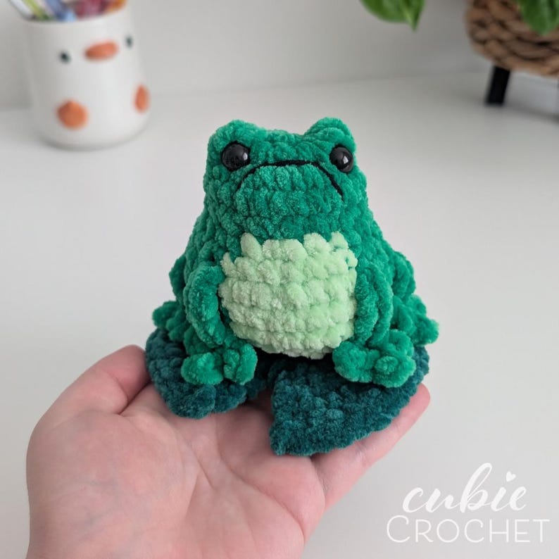 Op de afbeelding: Een kleine, zachte, donkergroene gehaakte kikker met een lichtgroene buik en zwarte plastic ogen. De kikker zit op een donkergroen gehaakt lelieblad. De tekst "cubie CROCHET" is zichtbaar in de rechterbenedenhoek van de afbeelding.