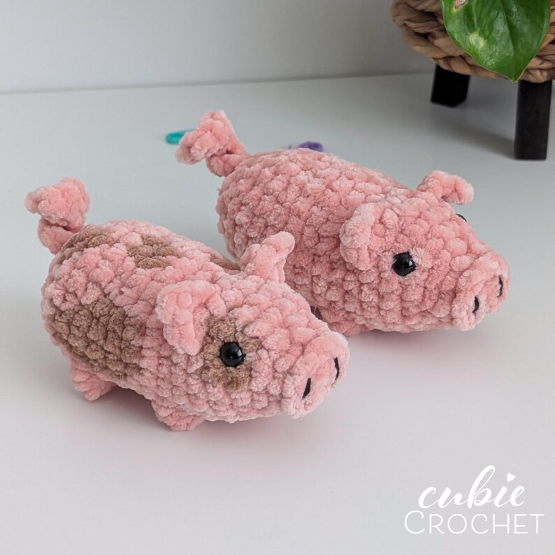 May include: Two handmade crochet pigs crafted from soft, plush pink yarn. One pig features light brown patches on its back and side. Both figures have small ears, curled tails, and black plastic eyes. The text cubie CROCHET appears in white.