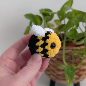 May include: A handmade, plush bee with a yellow and black body, white wings, and a black eye. The bee is round and fluffy, with a textured appearance. The background includes a green plant in a woven basket.