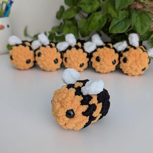 May include: A collection of handmade crochet bees. The bees are primarily orange and black, with white wings and black eyes. The plush toys are arranged on a white surface, with green foliage in the background.