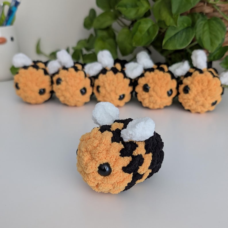 May include: A collection of handmade crochet bees. The bees are primarily orange and black, with white wings and black eyes. The plush toys are arranged on a white surface, with green foliage in the background.