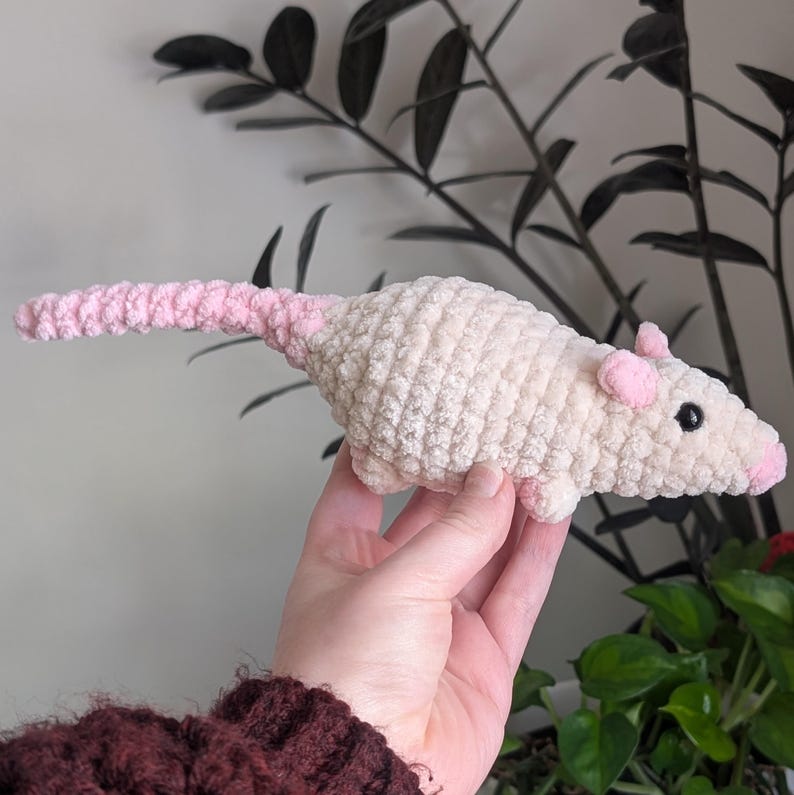 A hand holding up a warm off-white colored crochet rat plushie.