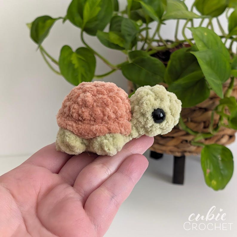May include: A small, handmade crochet turtle toy featuring a textured, dusty rose-colored shell and a light sage green head and body. The turtle has a single visible black plastic eye and a soft, plush yarn construction with a rounded shape.