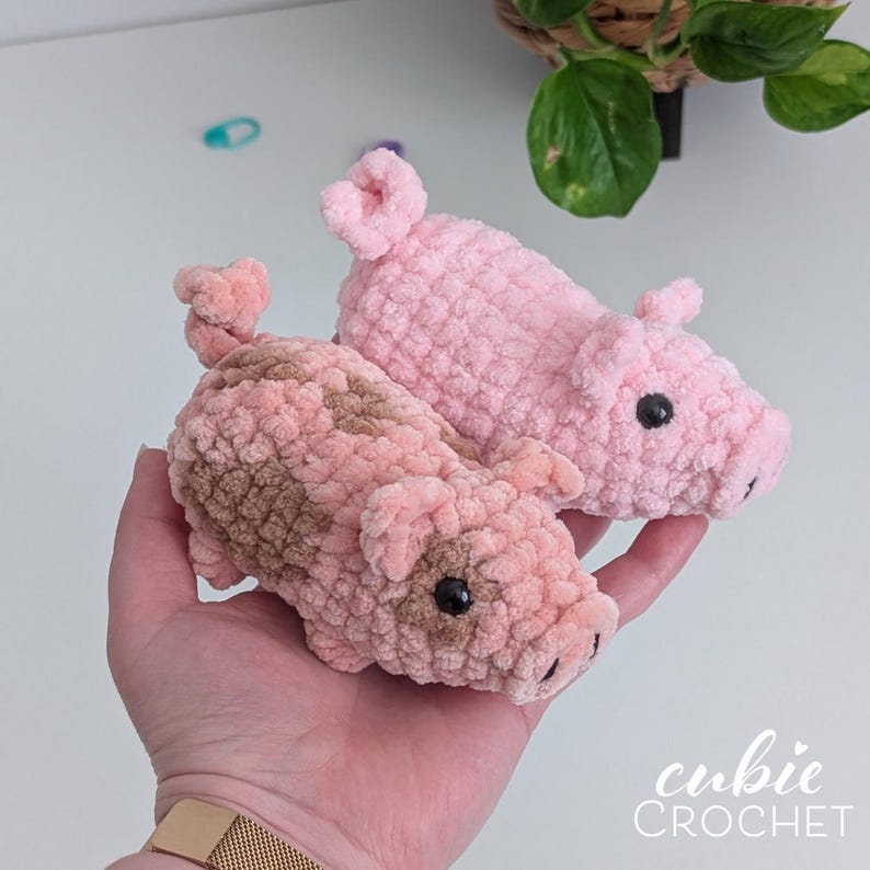 May include: Two small, plush, crocheted pig figures made from soft, textured yarn. One pig is solid light pink, while the other is a mix of light pink and brown patches. Both have small, curled tails, pointed ears, and black bead eyes.