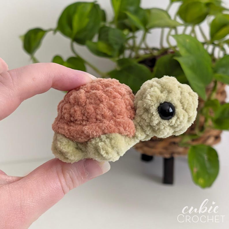 May include: A small, handmade crochet turtle plushie featuring a soft, sage green head and limbs with a textured, dusty rose-colored shell. The turtle has a single visible black plastic safety eye and is crafted from plush, fuzzy yarn.
