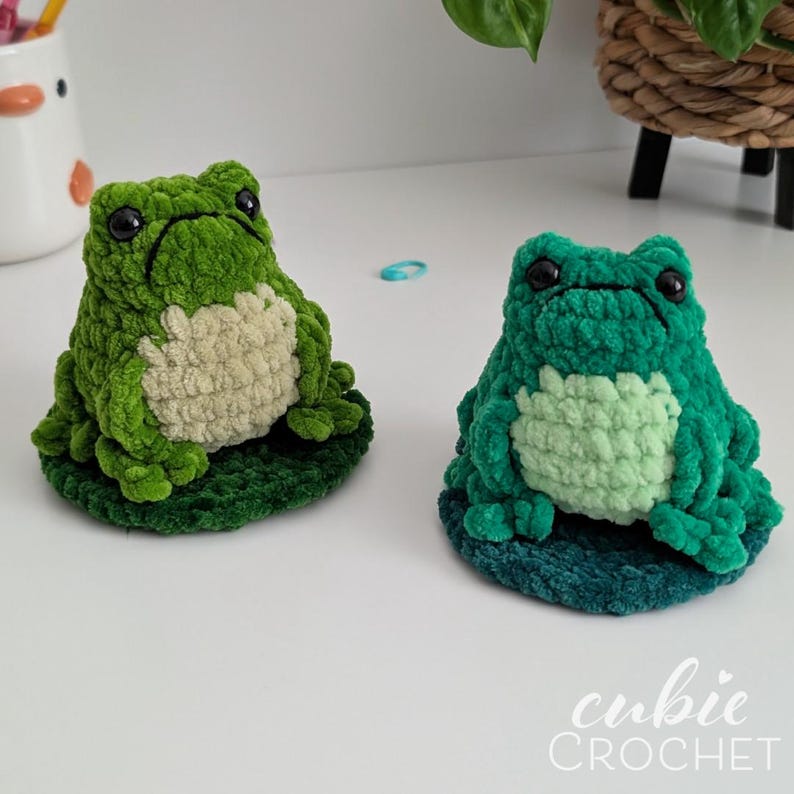 Op de afbeelding: Twee handgemaakte gehaakte kikkers van pluche groen garen. De linker kikker is olijfgroen met een cr&egrave;mekleurige buik, terwijl de rechter kikker donkerblauwgroen is met een lichtmuntkleurige buik. Beide hebben zwarte plastic ogen en zitten op ronde matjes.