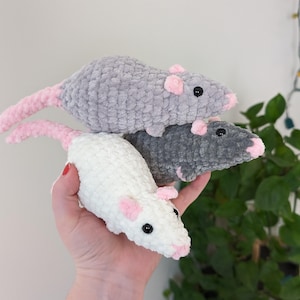 A hand holding up three crochet rat plushies. The one on the bottom is cream colored, the one in the middle is dark gray, and the one on the top is light gray. They all have black eyes and light pink noses, ears, and tails.