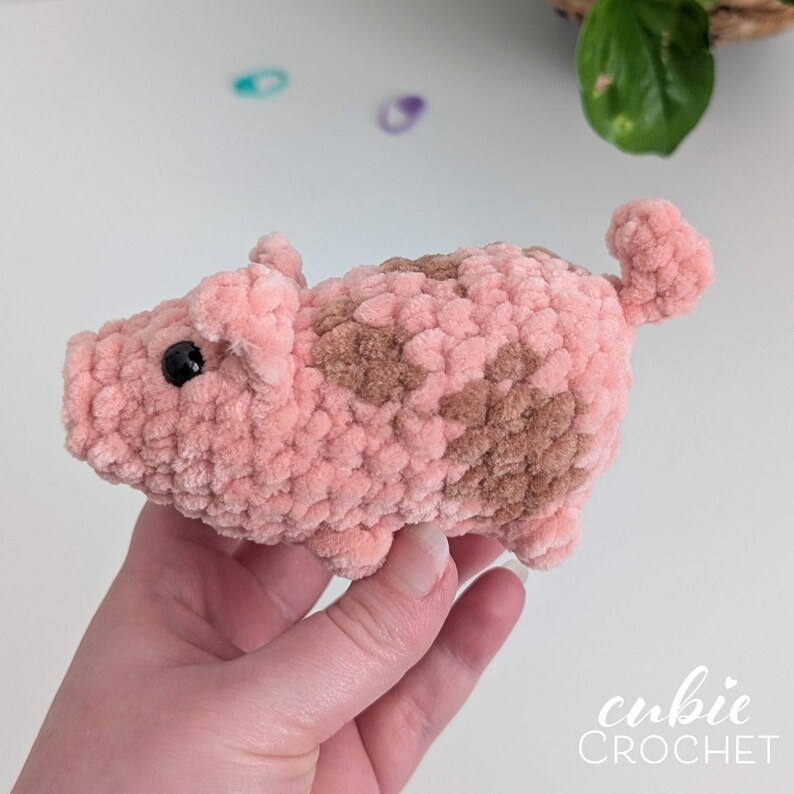 May include: A small, handmade crochet pig plush toy crafted from soft, textured pink yarn with light brown patches on its back. The pig features a curled tail, two small ears, and a black bead eye. The text cubie CROCHET is visible in the corner.