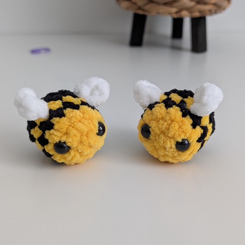 May include: Two handmade crochet bee toys. Each bee has a yellow body with black checkered stripes, black eyes, and white fuzzy wings. The bees are sitting on a white surface.