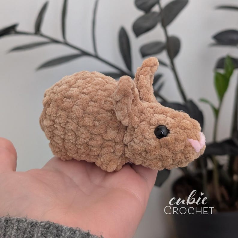 Op de afbeelding: Een handgemaakt, bruin gehaakt konijn met zwarte ogen en een roze neus. Het zachte speelgoed heeft een getextureerde afwerking. De tekst "cubic CROCHET" is rechtsonder zichtbaar. Een charmant, knuffelbaar speelgoed.