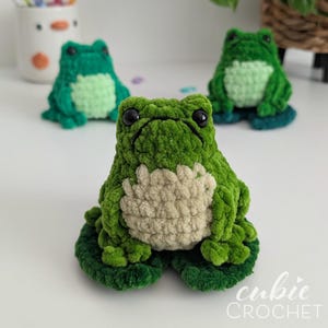 Op de afbeelding: Een kleine, handgemaakte gehaakte kikkerknuffel met een felgroen lichaam en een textuurrijke, lichtbeige buik. De kikker heeft twee grote, glanzende zwarte ogen en zit op een donkergroen gehaakt waterlelieblad. Het ontwerp is zacht en pluche.