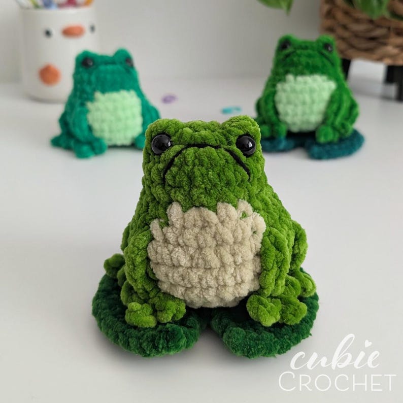Op de afbeelding: Een kleine, handgemaakte gehaakte kikkerknuffel met een felgroen lichaam en een textuurrijke, lichtbeige buik. De kikker heeft twee grote, glanzende zwarte ogen en zit op een donkergroen gehaakt waterlelieblad. Het ontwerp is zacht en pluche.