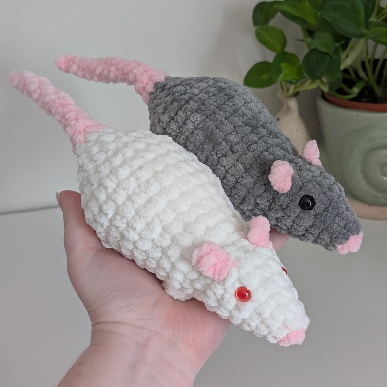 A hand holding up two crochet rat plushies. The one closer to the camera is a cream color with red eyes, and the other rat is dark gray with black eyes. They both have light pink noses, ears, and tails.