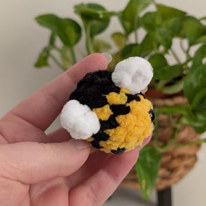 May include: A handmade, plush bee with a yellow and black body, white pom-pom wings, and black bead eyes. The bee is held in a hand, with a blurred background of green plants.