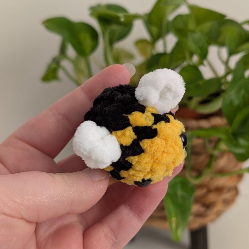 May include: A handmade, plush bee with a yellow and black body, white pom-pom wings, and black bead eyes. The bee is held in a hand, with a blurred background of green plants.