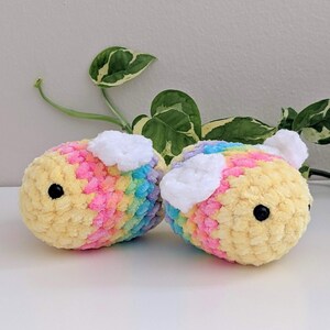 Puede incluir: Dos abejas de crochet de felpa hechas a mano. Las abejas son amarillas con rayas arco&iacute;ris rosas, azules y moradas. Cada abeja tiene un par de alas blancas y ojos negros. El fondo incluye plantas de hojas verdes y blancas.