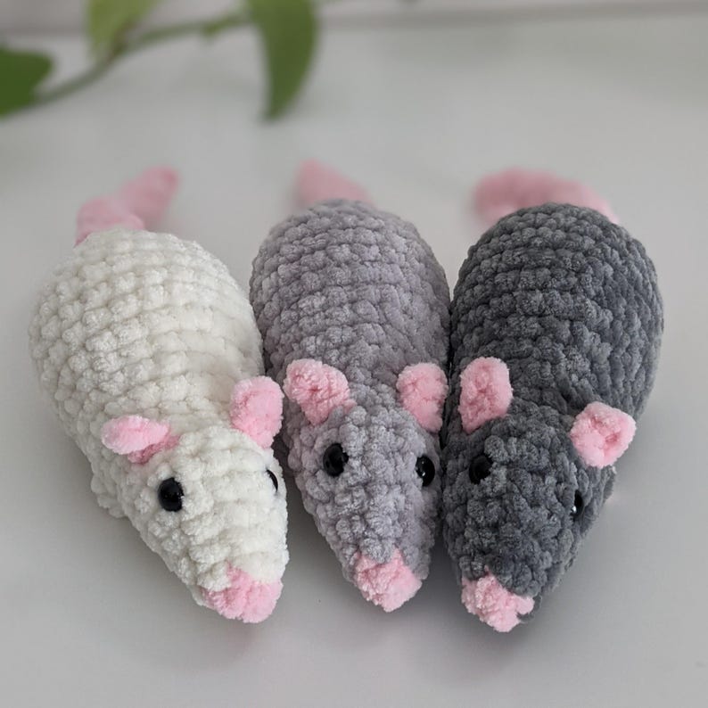 Three crocheted rat plushies, viewed from the front. Each one has a light pink nose, ears, and tail. The rat on the left is a cream color, the middle one is light gray, and the one on the right is dark gray.