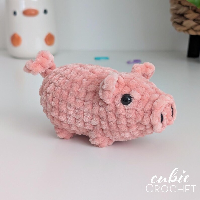 May include: A small, pink, plush pig made of soft, textured yarn. The pig features a curled tail, two small ears, and a snout with two dark nostrils. It has two round, black plastic eyes. The text "cubie CROCHET" is visible in the bottom right corner.