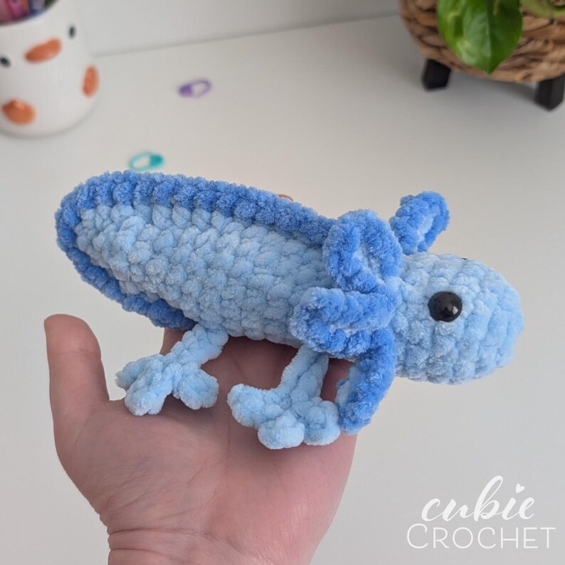 May include: A handmade blue crochet axolotl plush toy. The plush features a light blue body with darker blue accents on the gills, legs, and head. The axolotl has a black bead eye. The text "cubie CROCHET" is in the bottom right corner.