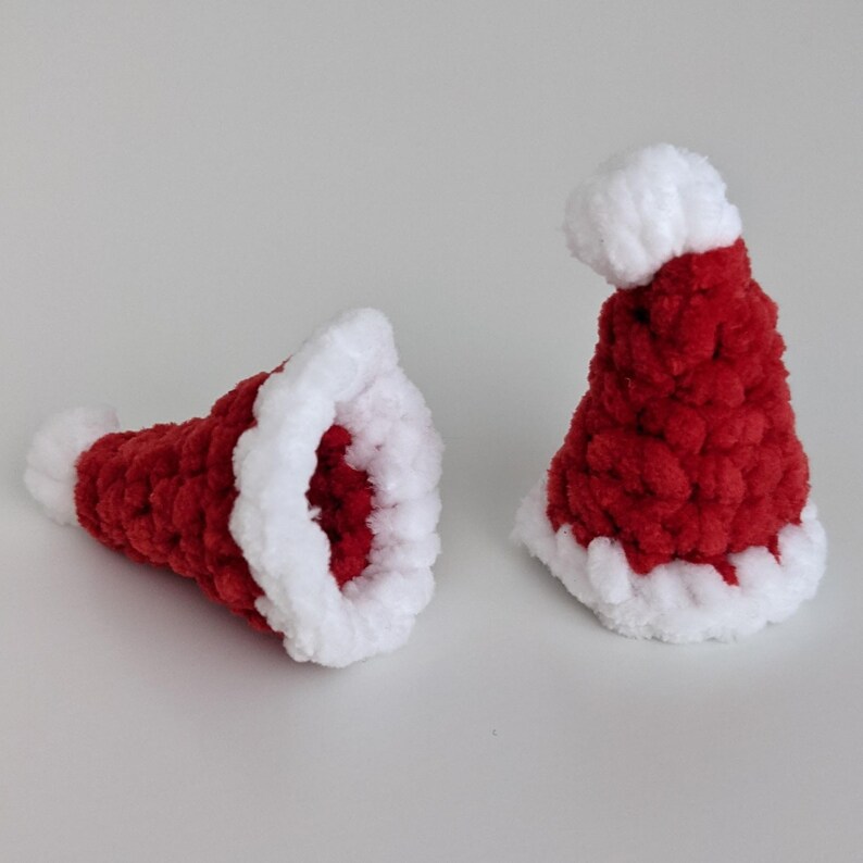 May include: Two miniature Santa hats. The hats are red with a white fluffy trim and a white pom-pom. The hats are made of a soft, textured material. One hat is upright, and the other is lying on its side.