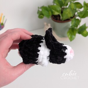 Op de afbeelding: Een handgemaakte gehaakte cavia in zwart en wit, met een roze neus en kleine zwarte ogen. Het handgemaakte knuffel is in een hand. Het "cubic CROCHET" logo is rechtsonder zichtbaar.