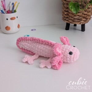 May include: A handmade, pink crochet axolotl toy with black eyes and detailed gills. The plushie is lying on a white surface. The text "cubie CROCHET" is in the bottom right corner. The axolotl is a popular aquatic salamander.