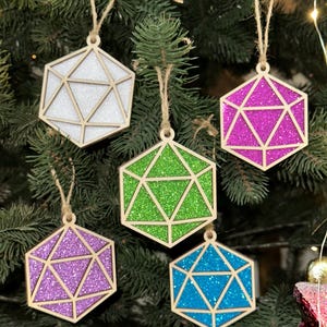 Op de afbeelding: Vijf geometrische kerstversieringen in witte, groene, paarse, blauwe en roze glitter, elk met een houten frame en touwhanger. De ornamenten hangen aan een groene kerstboom.