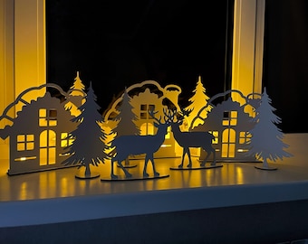 Puede incluir: Una escena invernal decorativa de madera con siluetas de casas, pinos y ciervos. Las casas tienen ventanas recortadas que brillan con luz cálida. La escena está sobre una superficie blanca.