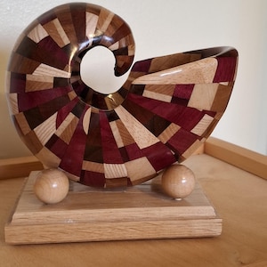 May include: A wooden nautilus shell sculpture, crafted from a mosaic of different wood tones including brown, red, and tan. The shell sits on a rectangular wooden base, supported by two small wooden spheres. A decorative item.