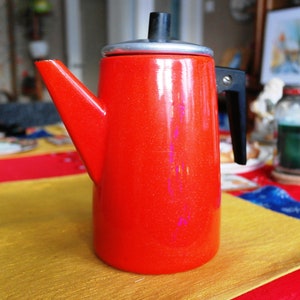 May include: A bright red coffee pot with a black handle and spout. The pot has a silver lid and a black top. The pot is on a yellow and red tablecloth. A cup and saucer are visible in the background.