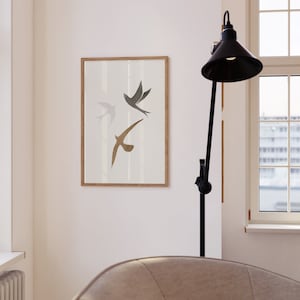 Op de afbeelding: Een ingelijste kunstdruk met drie gestileerde vogels in vlucht. De vogels zijn afgebeeld in grijstinten en bruin tegen een witte achtergrond. De lijst is lichtbruin. Het kunstwerk wordt aan een witte muur getoond.