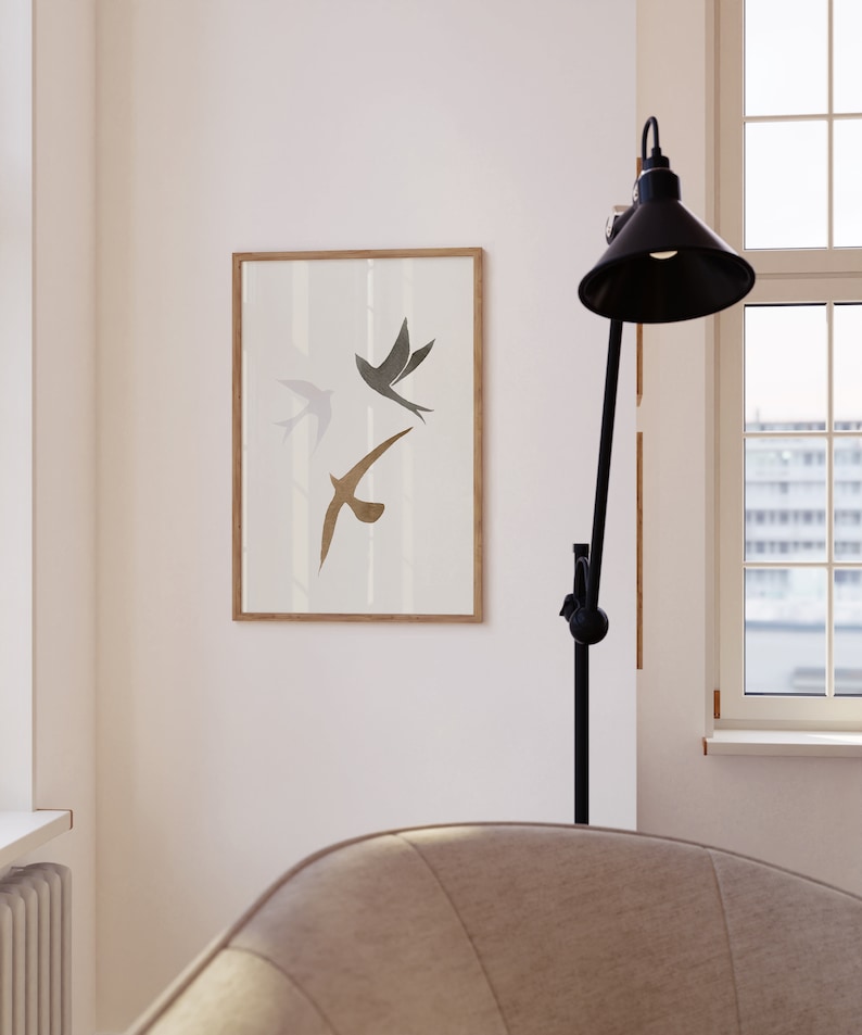 Op de afbeelding: Een ingelijste kunstdruk met drie gestileerde vogels in vlucht. De vogels zijn afgebeeld in grijstinten en bruin tegen een witte achtergrond. De lijst is lichtbruin. Het kunstwerk wordt aan een witte muur getoond.