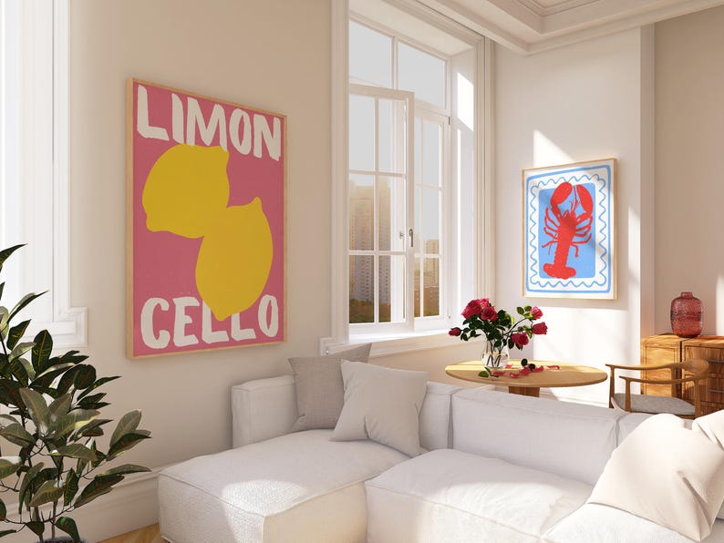 May include: Two framed art prints hang on a white wall. One features a pink background with yellow lemons and the words "LIMON CELLO." The other has a red lobster on a blue background with a white border. A white sectional sofa and round table are in the room.