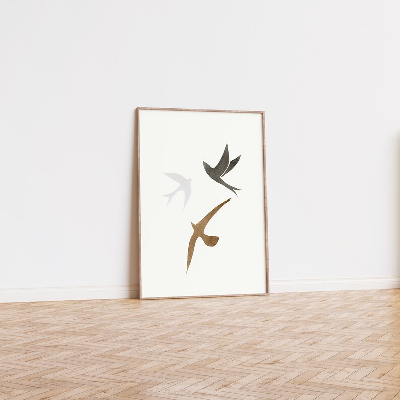 Op de afbeelding: Ingelijste kunstdruk met drie gestileerde vogels in vlucht. De vogels zijn afgebeeld in grijstinten en bruin tegen een cr&egrave;mekleurige achtergrond. De lijst is lichtbruin. Het kunstwerk leunt tegen een witte muur.