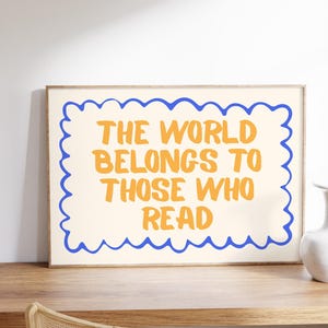May include: A framed print with the text "THE WORLD BELONGS TO THOSE WHO READ" in orange letters on an off-white background. The text is framed by a blue wavy border. The print is displayed on a wooden desk.