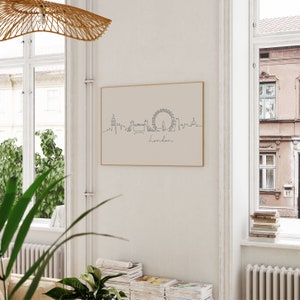 May include: A minimalist line drawing of the London skyline, featuring the London Eye, Big Ben, and other iconic landmarks. The drawing is printed on a beige background and framed in a thin, gold frame. The word "London" is written below the skyline.