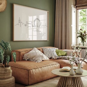 May include: A brown leather sectional sofa with white and black patterned pillows, a green pillow, and a throw blanket. The sofa is in front of a green wall with a framed print of the London skyline. A wooden coffee table with a book and two white vases with white flowers sits in front of the sofa.