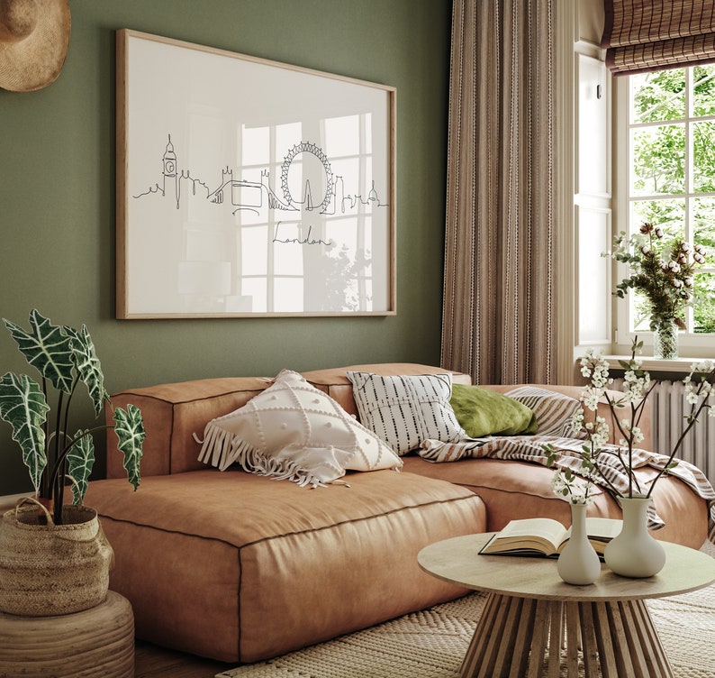 May include: A brown leather sectional sofa with white and black patterned pillows, a green pillow, and a throw blanket. The sofa is in front of a green wall with a framed print of the London skyline. A wooden coffee table with a book and two white vases with white flowers sits in front of the sofa.