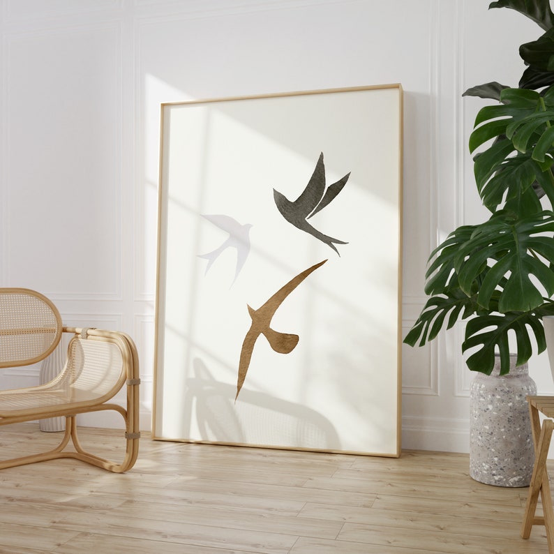 Op de afbeelding: Grote ingelijste kunstprint met drie gestileerde vogels in vlucht. De vogels zijn afgebeeld in grijstinten en bruin tegen een cr&egrave;mekleurige achtergrond. De lijst is licht van kleur en het kunstwerk wordt in een lichte kamer getoond.