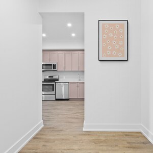 May include: A hallway with white walls and light wood floors. A doorway leads to a kitchen with pink cabinets, stainless steel appliances, and a white countertop. A framed print with a pink background and white flowers hangs on the wall.