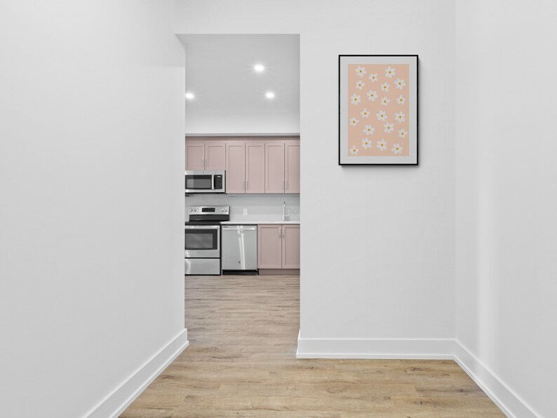 May include: A hallway with white walls and light wood floors. A doorway leads to a kitchen with pink cabinets, stainless steel appliances, and a white countertop. A framed print with a pink background and white flowers hangs on the wall.
