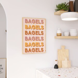 Puede incluir: Una impresi&oacute;n enmarcada con la palabra "Bagels" repetida seis veces en un dise&ntilde;o vertical apilado. El texto est&aacute; en una fuente retro con un esquema de color rosa, naranja y amarillo. La impresi&oacute;n est&aacute; enmarcada en un marco de madera claro y est&aacute; colgada en una pared blanca.