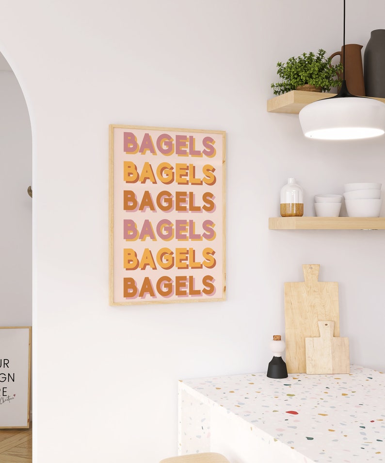 Puede incluir: Una impresi&oacute;n enmarcada con la palabra "Bagels" repetida seis veces en un dise&ntilde;o vertical apilado. El texto est&aacute; en una fuente retro con un esquema de color rosa, naranja y amarillo. La impresi&oacute;n est&aacute; enmarcada en un marco de madera claro y est&aacute; colgada en una pared blanca.