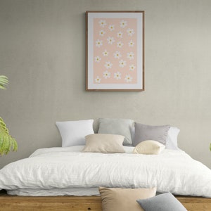May include: A white bed with a white linen duvet cover and a wooden headboard. There are several pillows on the bed, including a white pillow, a beige pillow, a gray pillow, and a white pillow with a floral pattern. A framed print with a pink background and white flowers hangs on the wall behind the bed.