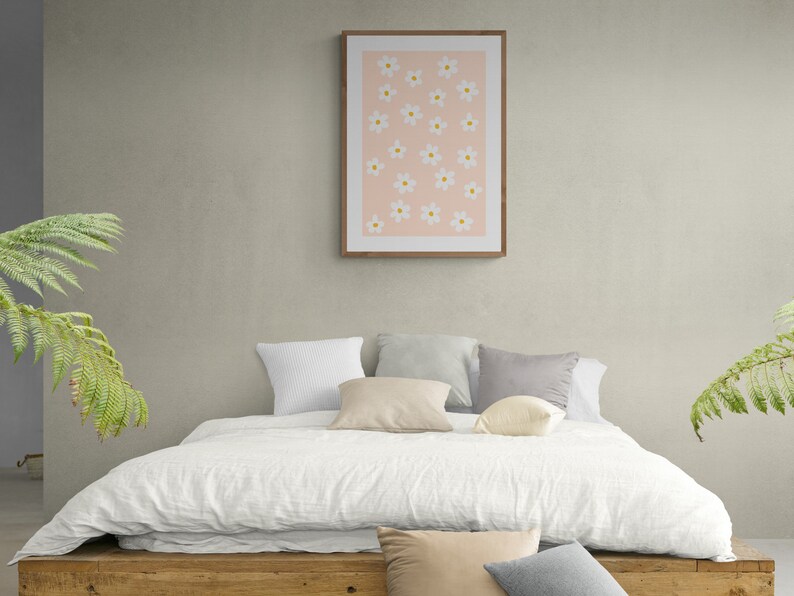 May include: A white bed with a white linen duvet cover and a wooden headboard. There are several pillows on the bed, including a white pillow, a beige pillow, a gray pillow, and a white pillow with a floral pattern. A framed print with a pink background and white flowers hangs on the wall behind the bed.