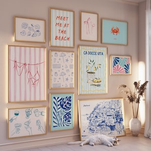 May include: A gallery wall with 10 framed prints featuring various designs, including a pink and white striped print with a bikini hanging on a clothesline, a blue and white print with lemons and the text "CA DOCCE VITA", a blue and white print with a stylised illustration of a cityscape, and a blue and white print with a stylised illustration of a crab.