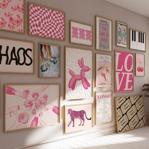 May include: A wall gallery featuring a variety of framed prints in pink, black, and white. The prints include abstract designs, text, and illustrations. The prints are arranged in a grid pattern on a light gray wall.